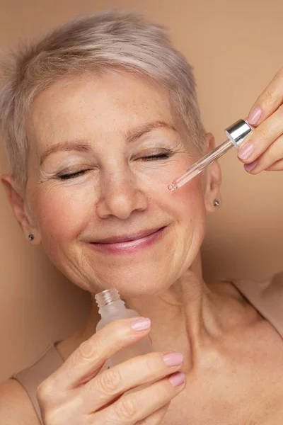 Happy senior lady applies cosmetic oil serum on face takes care of skin and smiles broadly enjoys beauty treatments stays always young beautifull poses against beige background