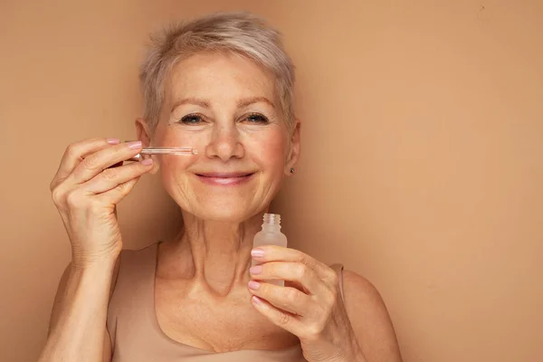Happy senior lady applies cosmetic oil serum on face takes care of skin and smiles broadly enjoys beauty treatments stays always young beautifull poses against beige background