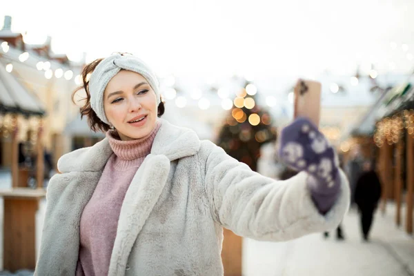 Charming young woman in winter clothes at the Christmas market takes a selfie on a smartphone, winter time