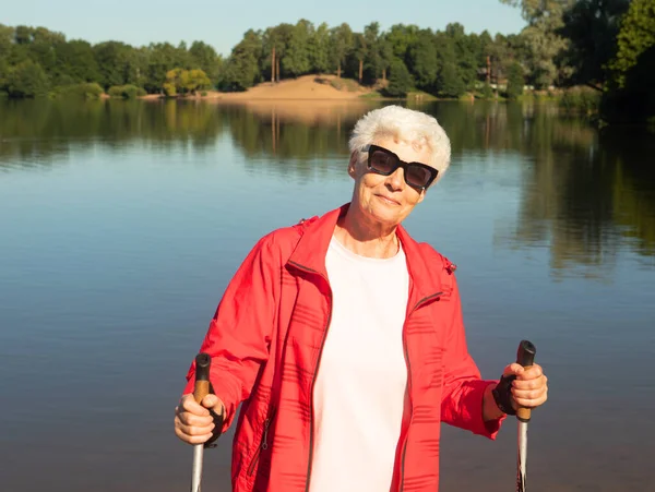 一个戴太阳镜的白发女人带着跟踪棒在湖边散步 健康的生活方式和快乐的老年观念 — 图库照片