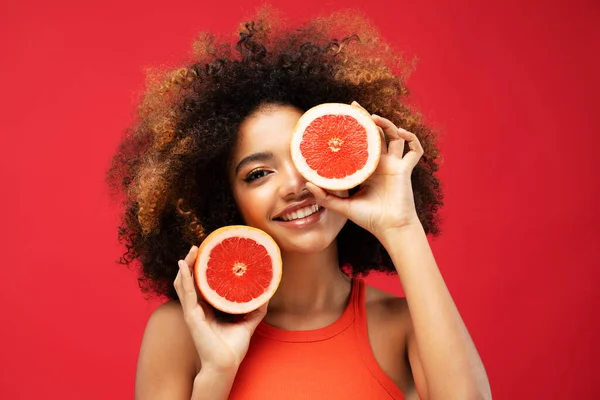 lifestyle, food, diet and people concept: Photo of young cheerful dark skin woman hold grapefruit cover eye look empty space isolated on red color background