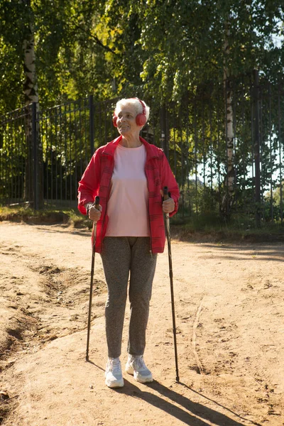 积极的生活方式 休闲和业余爱好的概念 身穿红色夹克 头戴耳机 精力充沛的健康老年女性在户外拍摄 在阳光明媚的海滩上使用北欧杆 — 图库照片