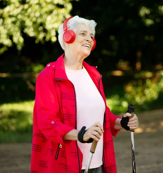 一个白发苍苍 留着短发 身穿红色夹克的漂亮老妇人 走在人行道上 脚踏着手杖 听着音乐 夏天的一天健康生活方式的概念 — 图库照片