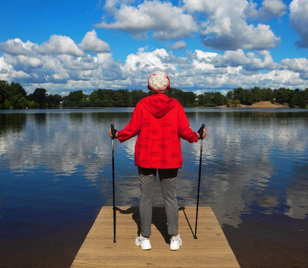 积极的生活方式 休闲和业余爱好 一个身穿红色西装的精力充沛的老妇人站在湖边的快照 一个女人拿着远足的杆子 欣赏着云彩 从后面看 — 图库照片