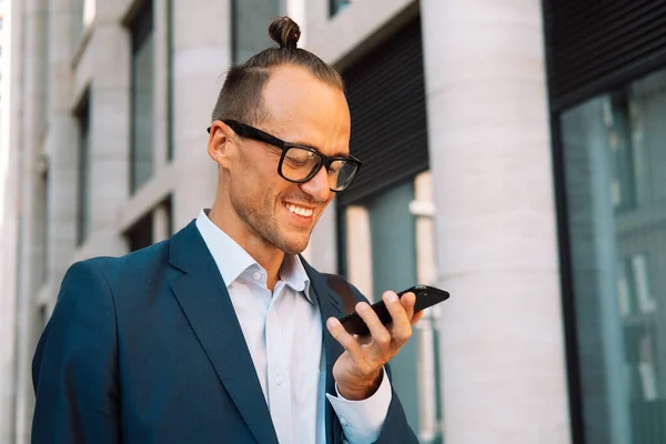一个英俊的年轻人在城市街道上走的时候 戴着眼镜 在智能手机上录音留言 男人在户外用智能手机传送语音讯息 — 图库照片