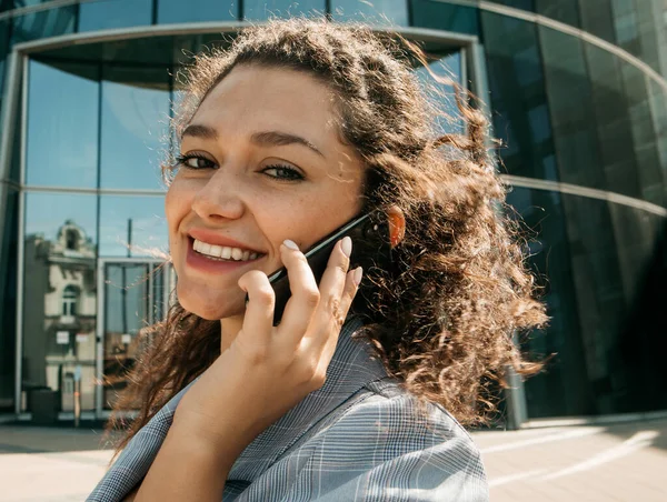 A young woman dressed in business style against the background of a business center is holding a mobile phone and talking.