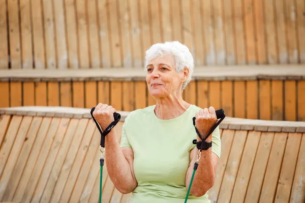 老妇人穿着绿色的T恤衫在户外做运动 坐在木制长椅上使用阻力橡皮筋 健康的生活方式 积极的退休生活和运动时间 — 图库照片