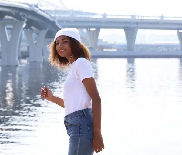 迷人的年轻非洲裔美国女人戴着白色的帽子 在靠近大海的地方用耳机听音乐 生活方式概念 — 图库照片