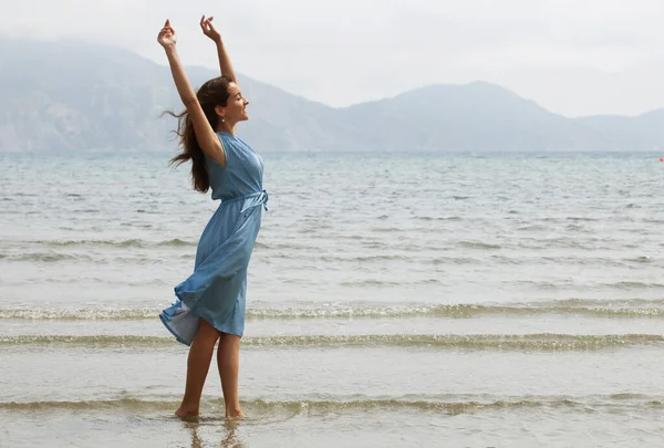 青いドレスを着た若いブルネットの女性がビーチを裸足で歩き 水の中で足をぶら下げています 夏休み明けましておめでとう — ストック写真