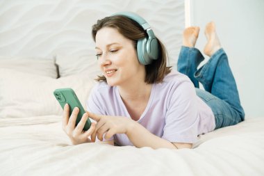 Gülümseyen yetişkin kadın kulaklıklarla yatağa uzanıyor ve elinde akıllı bir telefonla raylara bakarken müzik dinliyor.