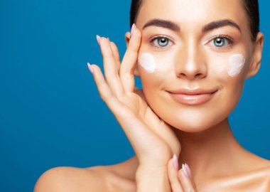 Yüzüne kozmetik krem süren temiz ciltli güzel genç bir kadın mavi arka planda izole edilmiş.