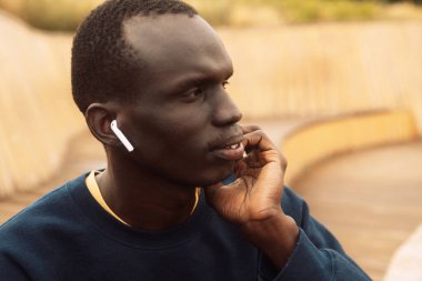 Koyu tenli genç bir adam parktaki ahşap bir bankta oturur ve müzik dinler. Yaşam tarzı ve insan kavramı.