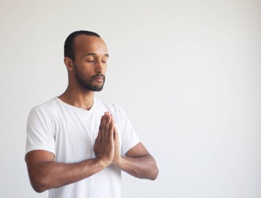 Beyaz tişört giyen genç afro adam, zihnine konsantre oluyor, el ele tutuşuyor, meditasyon yapıyor, yoga yapıyor, nefes egzersizi yapıyor stresi azaltıyor..