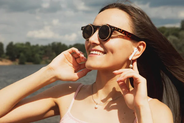 Cheerful happy young woman with long hair in sunglasses listening to music next to the lake. Happy summer. Lifestyle concept.