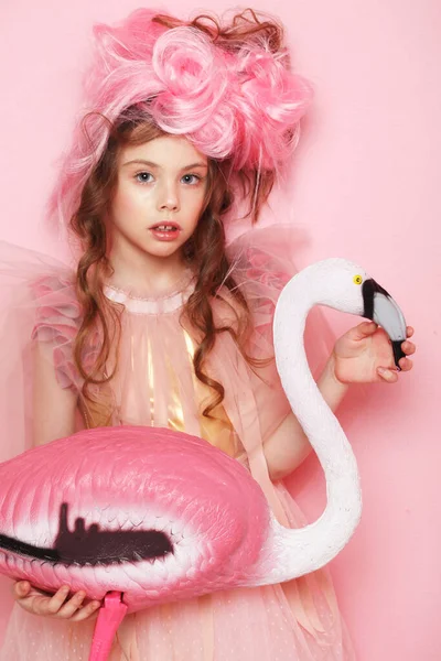 Beautiful little child girl in pink dress with bright make-up holding a flamingo bird over pink background