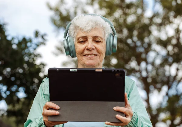 タブレット付きヘッドフォンで祖母は音楽を聞きます 公園のベンチに座って音楽を聴くタブレット付きのヘッドフォンの高齢者女性 — ストック写真