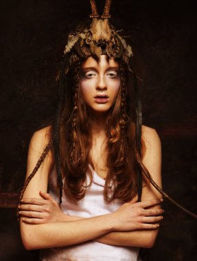 Parlak makyajlı, örgülü ve kafasında kafatası olan genç bir kadın kollarını siyah bir arka planda kavuşturmuş duruyor.