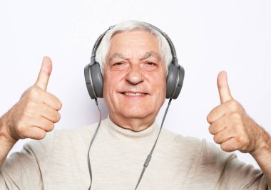 Gündelik kıyafetleri ve kulaklıkları olan yakışıklı yaşlı adam, beyaz arka planda cep telefonu ve gülümseme kullanarak müzik dinliyor.