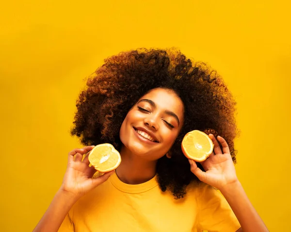 lifestyle, food, diet and people concept: Charming dark-skinned young woman in a yellow t-shirt holds orange slices. Beautiful model posing with her eyes closed, on a yellow studio background.