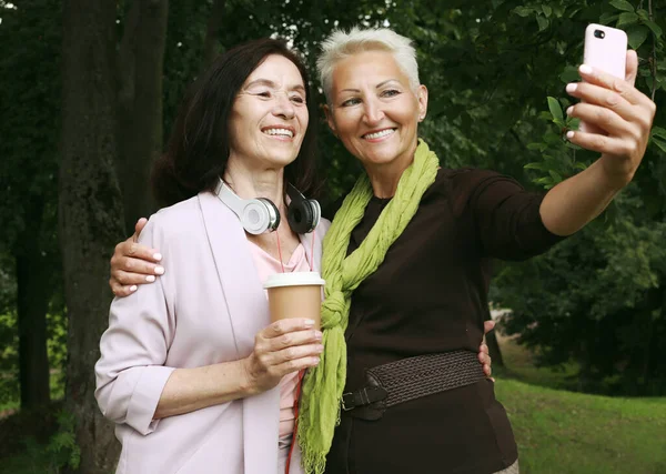 Two charming elderly women friends holds a cup of coffee to go and making selfie in summer park. Lifestyle and gold age concpet.