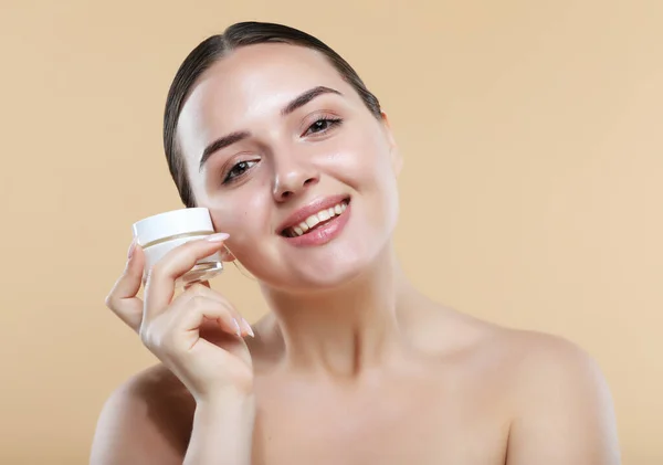 Beauty Youth Skin Care Concept - Beautiful Caucasian Woman Face Portrait holding and presenting cream jar product. Beautiful Spa model Girl with Perfect Fresh Clean Skin over beige background.