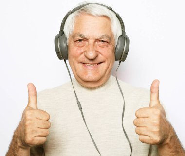 Yaşam tarzı, tehnoloji ve yaşlılar konsepti: Sıradan giyinen ve kulaklık takan yakışıklı yaşlı adam müzik dinliyor ve beyaz arka planda OK işareti gösteriyor