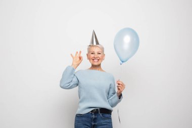 Güzel görünümlü, çekici, neşeli, neşeli, neşeli, gri saçlı, elinde mavi balon olan kadının portresi. Tamam, beyaz gri arka planda izole edilmiş.