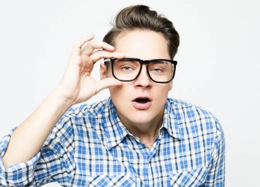 Mavi günlük ekose gömlek ve gözlük giyen genç adam beyaz arka planda heyecanlı bir yüz görünce şaşırdı ve şok oldu.