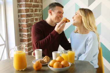 Yaşam tarzı, yemek, aile ve insan konsepti: mutfakta kahvaltı yapan sevimli genç bir çift, kadını kruvasanla besleyen erkek, mutlu zaman..
