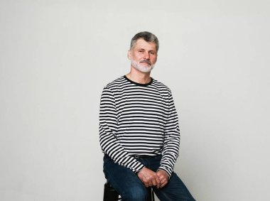 Yaşam tarzı, duygu ve yaşlı insanlar konsepti. Çizgili hırkalı yaşlı sakallı adam gri bir stüdyo arka planında bir sandalyede oturuyor.