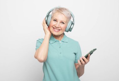 İnsanlar, teknoloji, yaşlanma ve olgunluk kavramı. Gri arka planda izole edilmiş kulaklıklı ve cep telefonlu güzel beyaz kadın fotoğrafı.