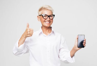 Modern yaşam tarzı, yaşlılar, iletişim ve elektronik aletler. Peri sarısı saçlı çekici yaşlı kadın açık gri arka planda yeni bir akıllı telefon sunar..