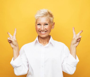 Portrait of happy and charming european senior woman with blond short hair laughing and show v-sign over yellow background