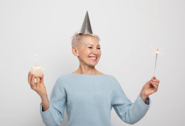 Happy middle aged female wearing conical hat celebrating birthday with cupcake with candle and sparkler over grey background