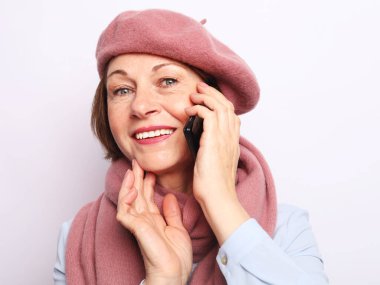 Beyaz gri arka planda pembe fular takan ve akıllı telefonlu şapka takan yaşlı bir kadın..