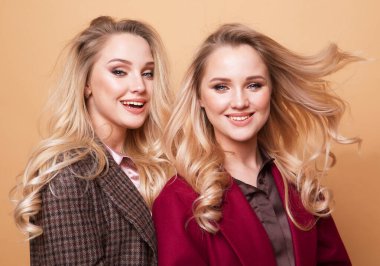 Moda ve güzellik konsepti: Bej arka planda poz veren, gündelik elbiseli iki güzel genç kadın. Moda bahar yaz fotoğrafı, güzel saçlar, büyük bir gülümseme. Arkadaşlık. İki kız kardeş.