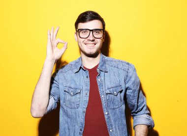 Basit giyinmiş yakışıklı, heyecanlı bir adamın portresi gülümseyen ve sarı arka planda izole edilmiş kameraya ok işareti gösteren.