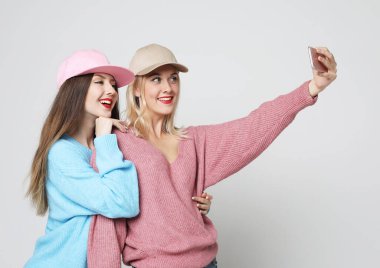 Portrait of two female friends dressed in sweaters and caps standing together and taking a selfie over grey background