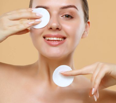 Stüdyodaki bej rengi arka planda pamuk pedi olan çekici, genç bir kadın temizlik yüzü. Portreyi kapat.