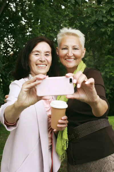 Two charming senior women friends holds a cup of coffee to go and making selfie in summer park. Lifestyle, friendship and gold age concpet.