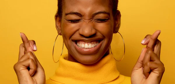 若いアフリカ系アメリカ人女性の肖像画は奇跡を待っています 祈りと希望の夢が叶う 幸運のための指を渡ります 目を閉じます 広く笑顔 黄色のジャンパーを身に着けています — ストック写真
