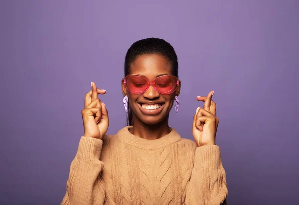 人々のコンセプト ピンクのサングラスの中で喜びの希望とカリスマ楽観的なアフリカ系アメリカ人のスタイリッシュな女性夢のためのクロス指笑顔紫の壁の上に真来る — ストック写真