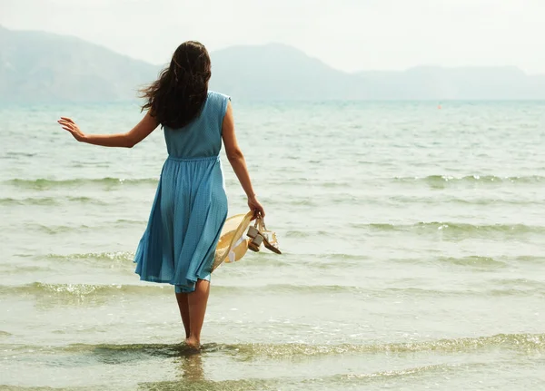 青いドレスを着た若いブルネットの女性が裸足でビーチを歩いていて 水の中で足をぶら下げているイメージ バックビュー — ストック写真