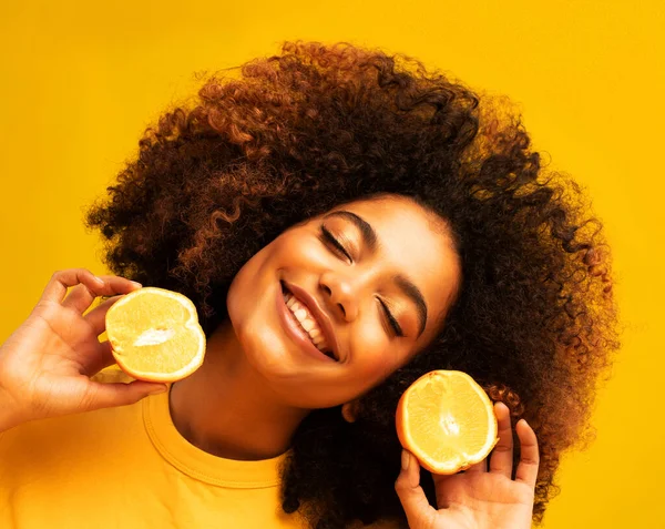 lifestyle, food, diet and people concept: Optimist Young female model of afro appearance. Clean, even skin of the face. In the hands of citrus, orange. Photo on yellow background.