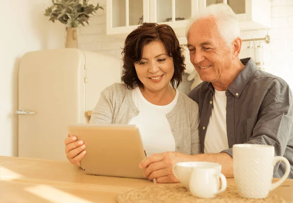 高齢者のカップルは キッチンに座って タブレットを使用して ニュースを見て 一緒に過ごす時間を楽しんでいます 現代の年金受給者はガジェットを使用 — ストック写真