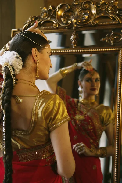 Female model Hindu Bride in saree, wearing gold and jasmine flower garlands in the hair near mirror, back view.