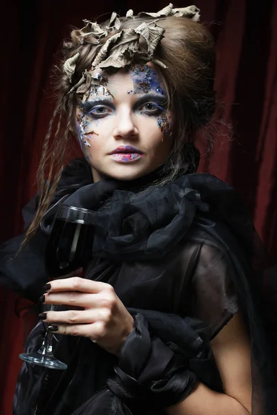 Party, holiday and Halloween concept. A beautiful woman in a witch costume with bright makeup and curly hair is holding a glass of wine.