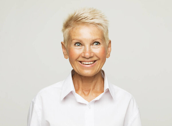 Жизнь начинается только с возрастом. Портрет счастливой и очаровательной европейской пожилой женщины с короткими светлыми волосами в белой рубашке на сером фоне.