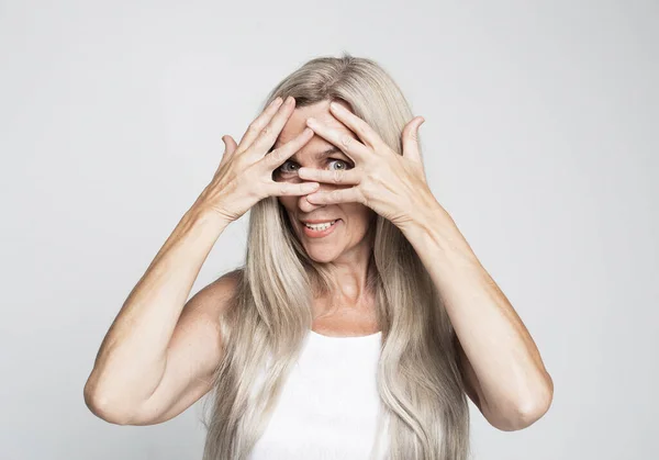 Smiling mature woman 60 years old with long hair closes her eyes with her palm on grey color background