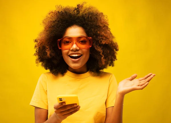 Excited cheerful dark skinned curly young woman raises palm and stares overjoyed at smartphone display being social media blogger reads amusing article or watches funny video communicates online